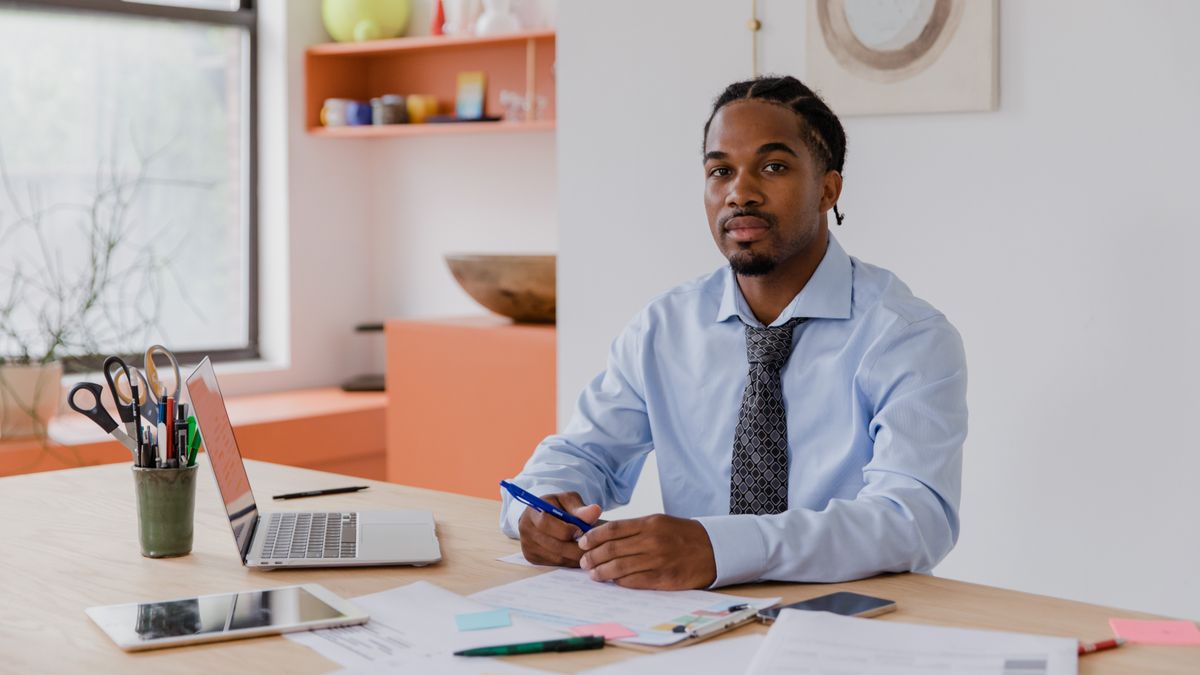 A professional businessman with a beard and braided hair sits at his desk with a laptop and documents, working diligentl