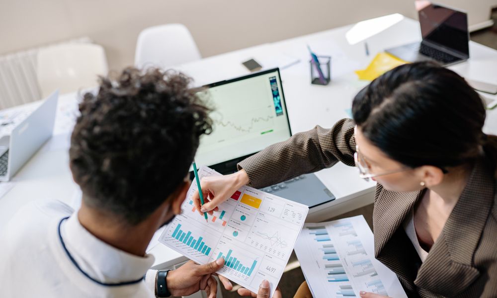 Two colleagues in a meeting room discussing financial charts and graphs on a laptop and paper.