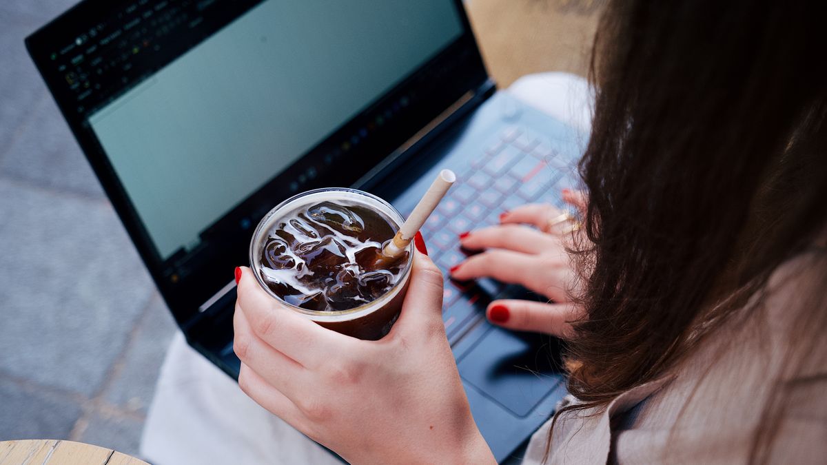 An adult woman working on a laptop while holding an iced drink, seated outside.