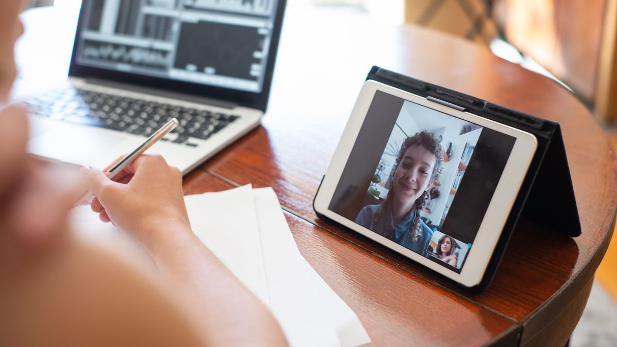 A person on a video call using a tablet, with a laptop displaying data nearby.