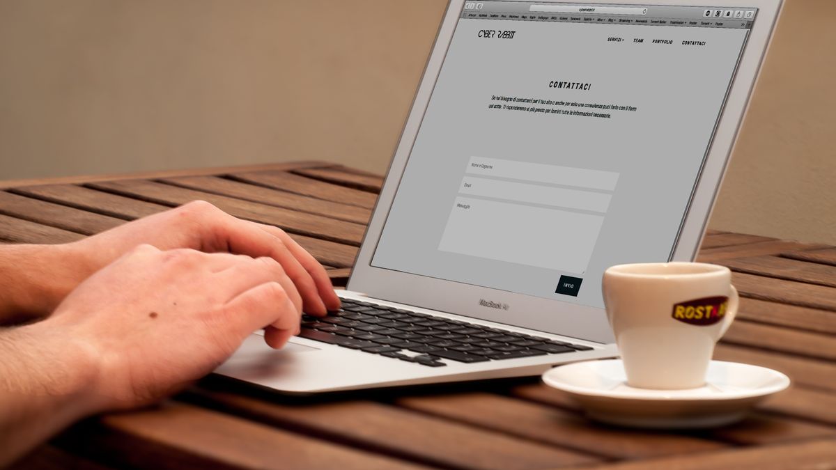 Close-up of hands typing on a laptop next to a cup of coffee on a wooden desk.