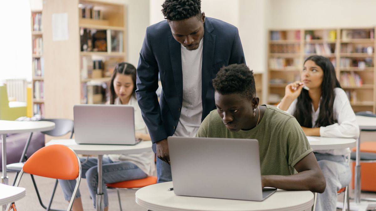 Students learning in a classroom setting with a teacher assisting and laptops on desks, creating an interactive educatio