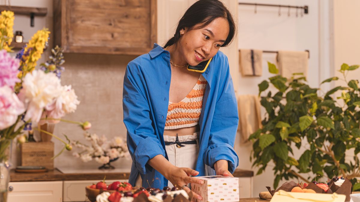 Asian woman in blue shirt and crop top packing cupcakes at a bakery, smiling while on a phone call.