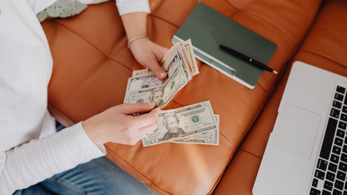 Person counting dollar bills on a sofa next to a notebook and laptop.
