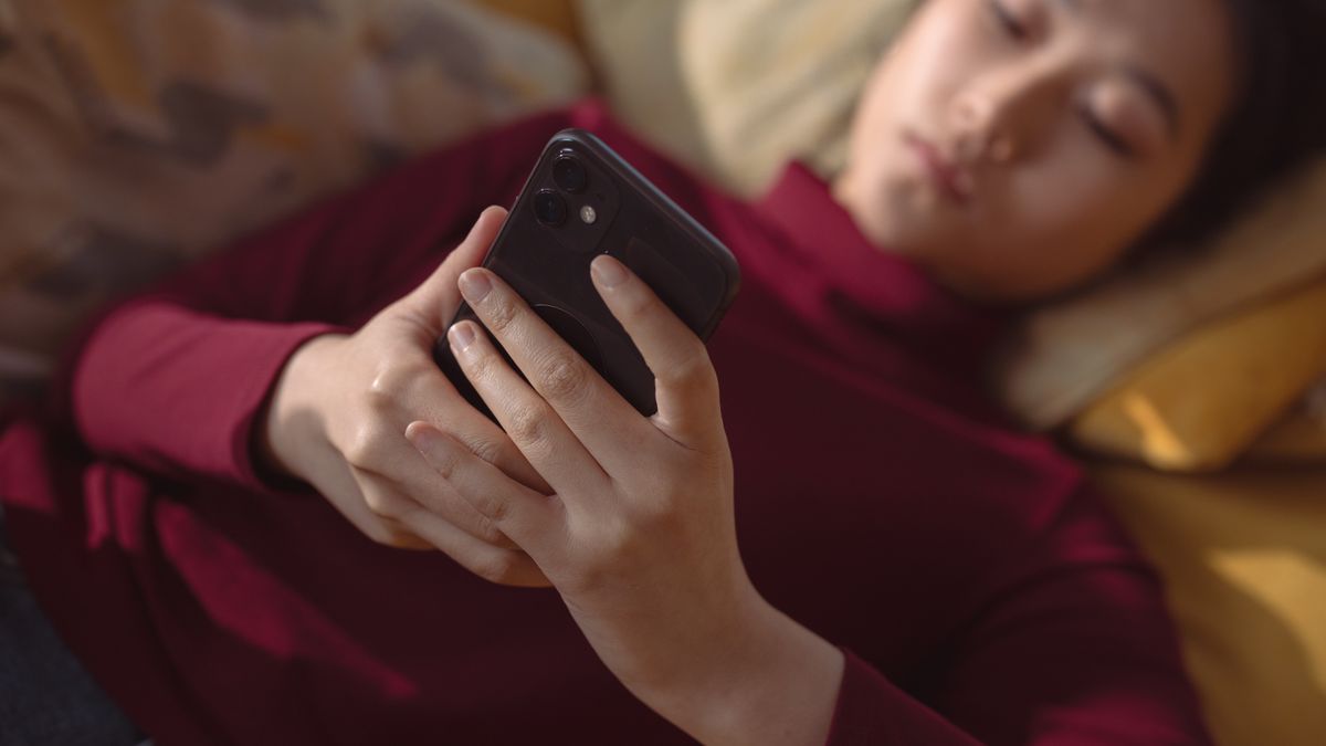 A young woman lying down, holding and using a smartphone for relaxation indoors.