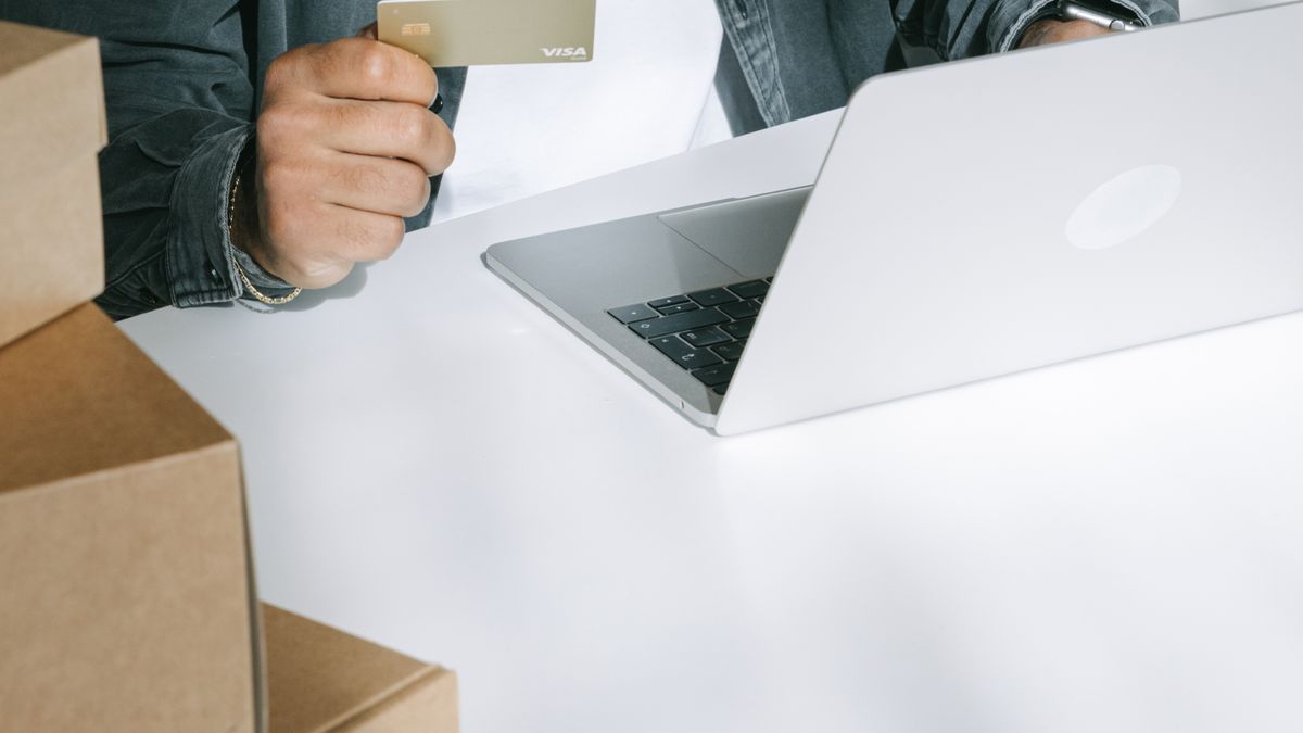 Person using a credit card for online shopping on a laptop surrounded by shipping boxes.