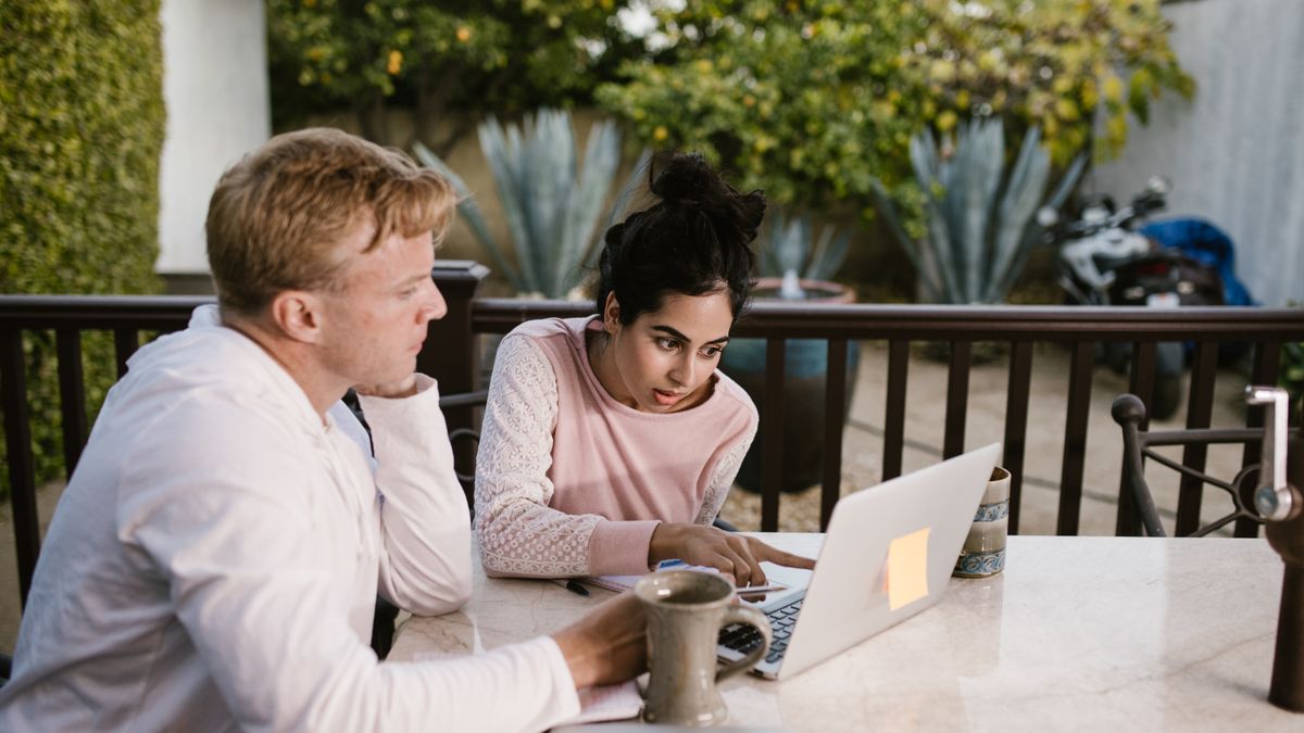 Two adults collaborating on a laptop, seated outdoors with coffee on the table.