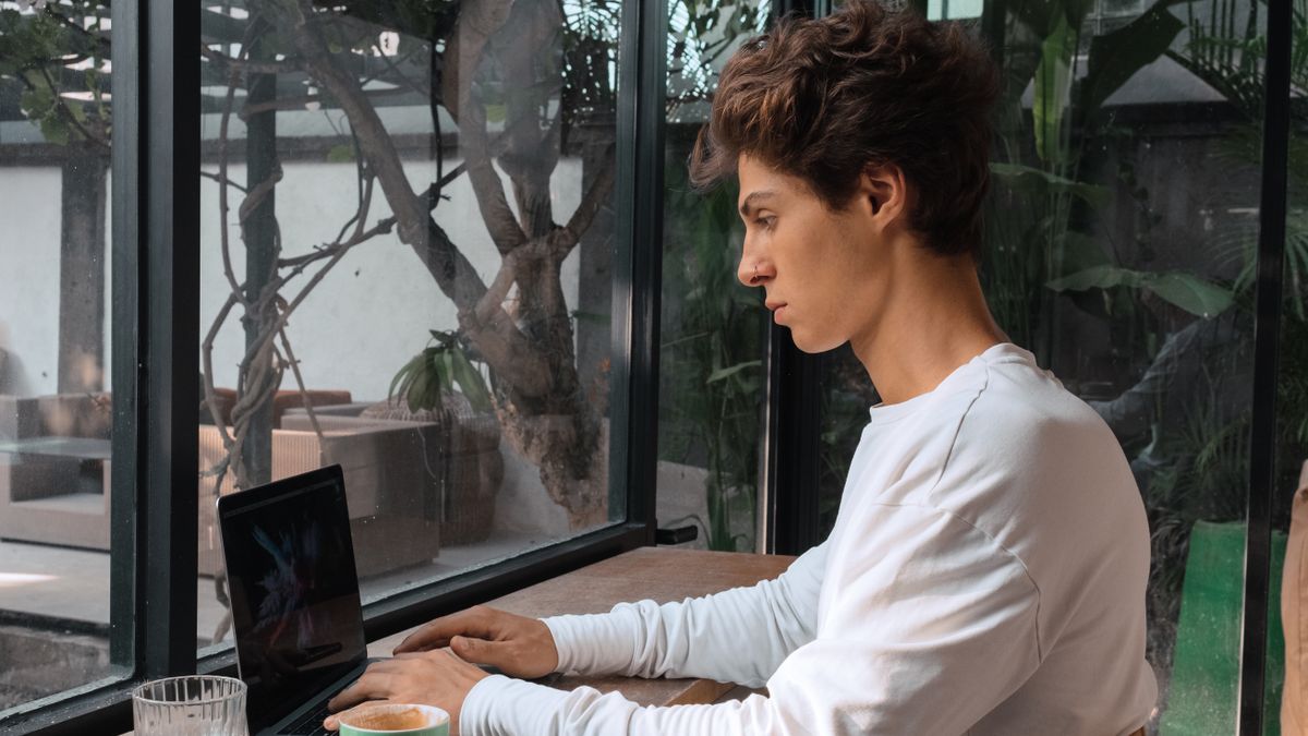 Focused young man typing on a laptop inside a cozy café, enjoying coffee by the window.