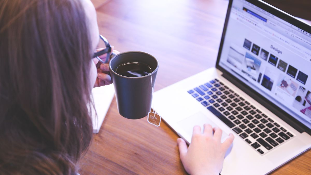 A woman enjoys coffee while browsing the web on a laptop at a home office desk.