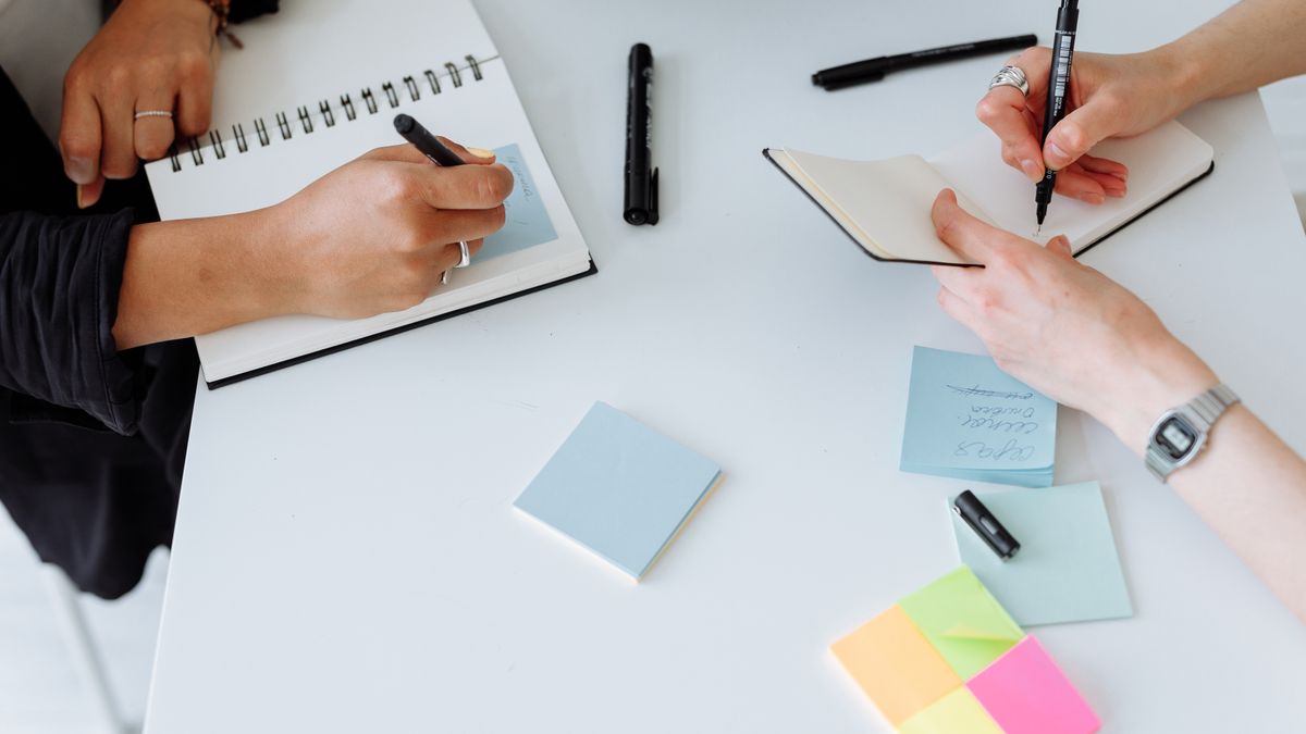 Top view of people collaboratively writing notes on a desk with notebooks and sticky notes.