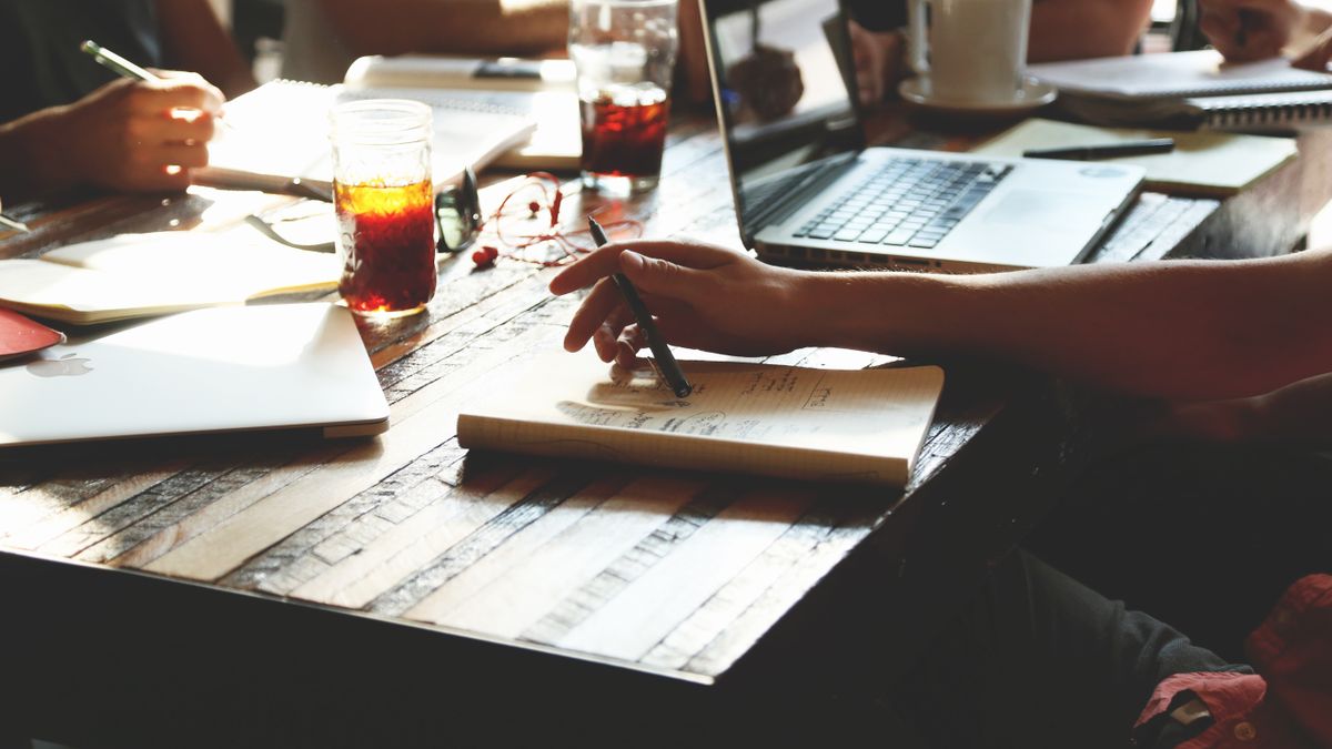 A group of adults engaged in a collaborative office meeting, discussing ideas with notes, laptops, and refreshments.