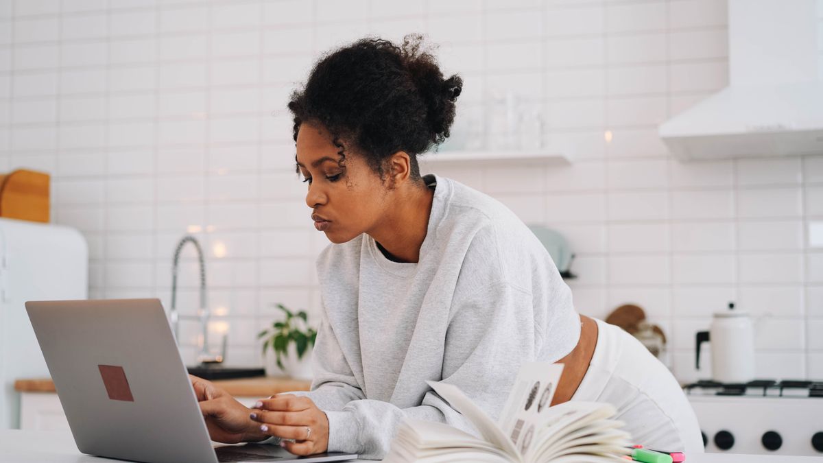 Woman at home in kitchen using laptop for online education with books around.