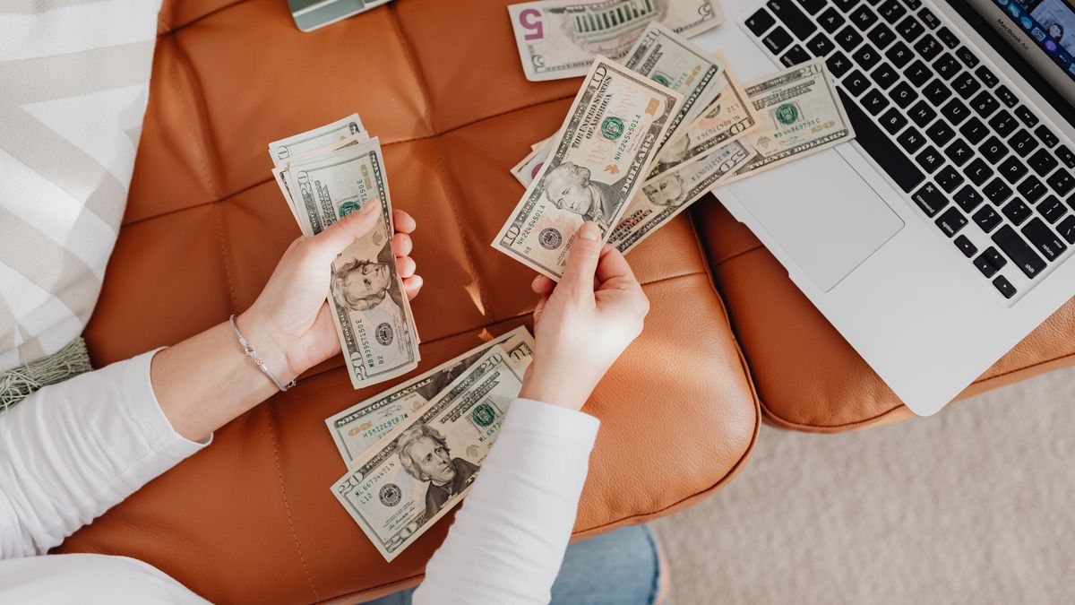 A person counts US dollar bills on a couch with a laptop, symbolizing financial planning.