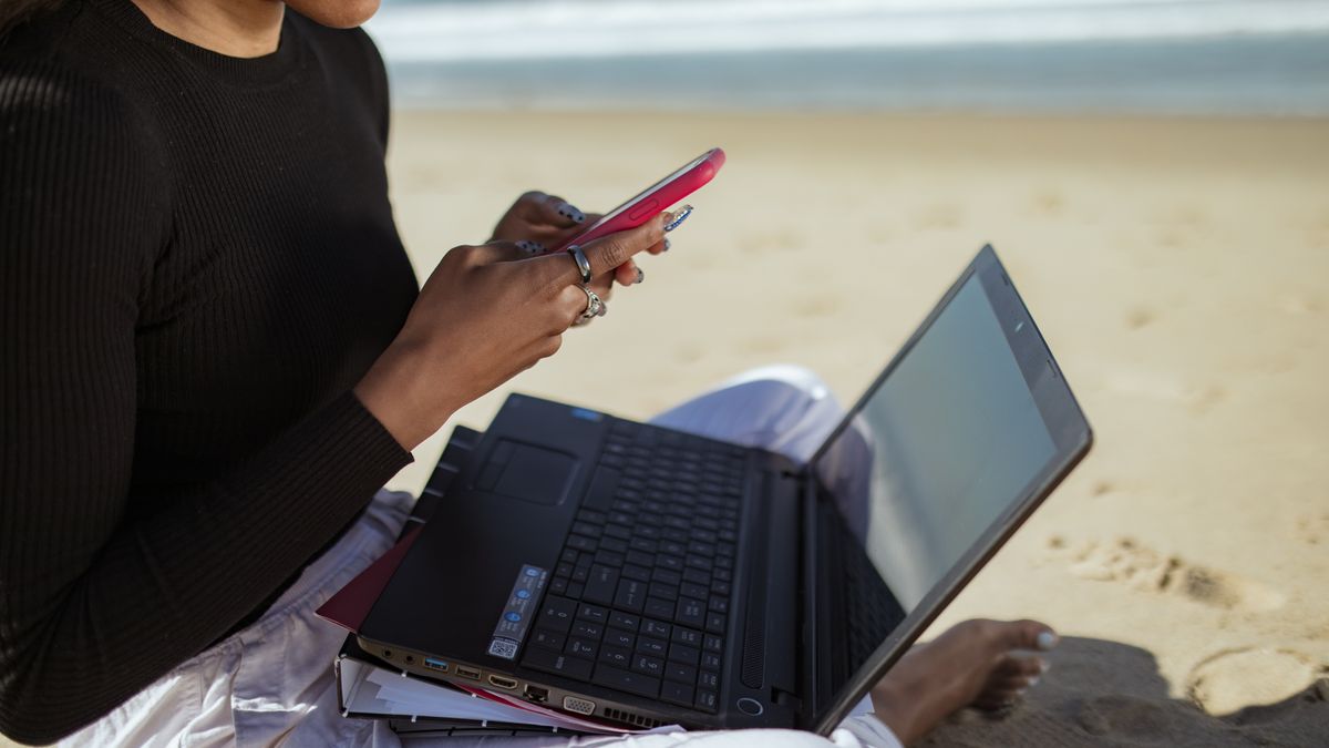 Person multitasking with a laptop and smartphone by the beach, embracing remote work lifestyle.
