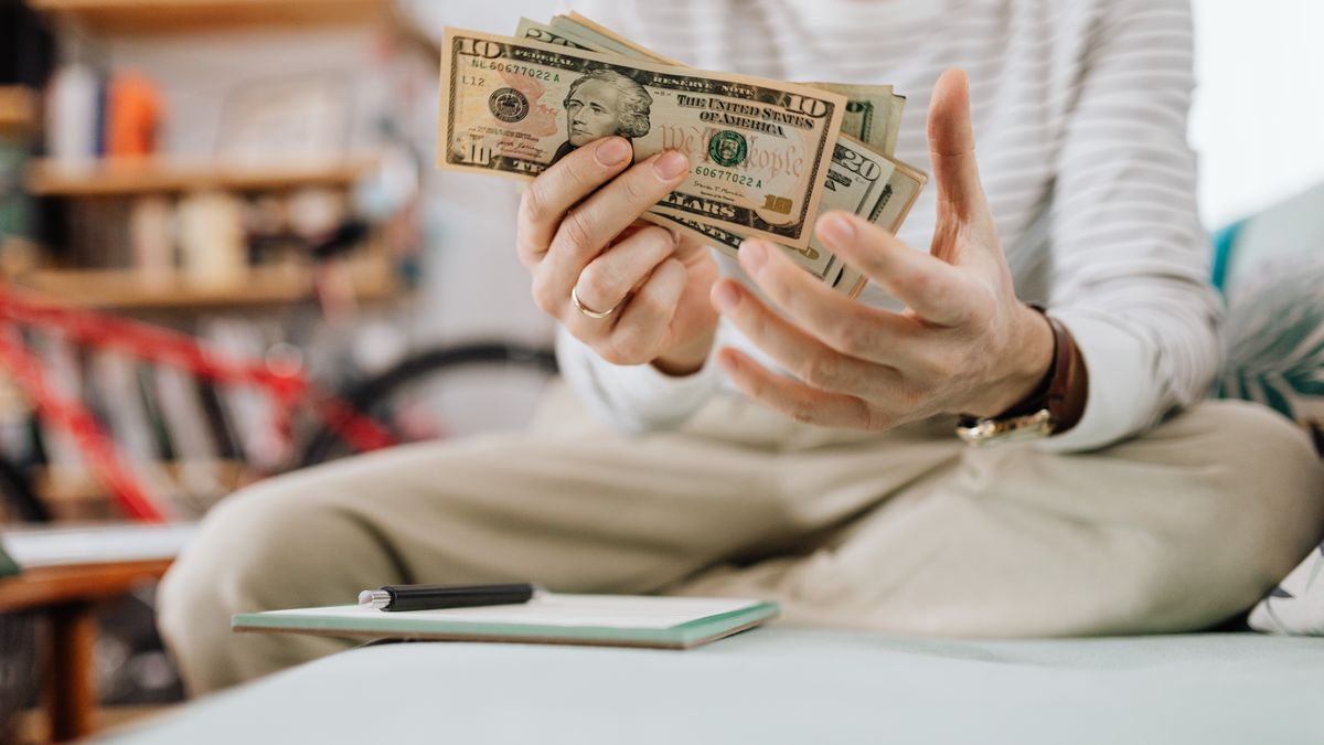 Man wearing casual clothes counts dollar bills while sitting on a sofa indoors.